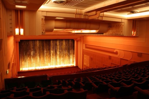 A cinema auditorium with warm wood-paneled walls and rows of seats facing the screen