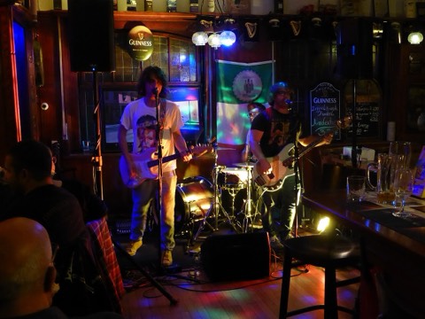 A band playing guitars and drums on a small stage in a crowded, dimly lit pub