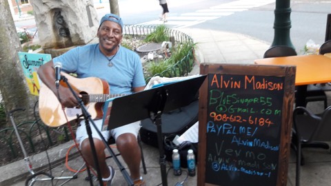 A singer-songwriter playing acoustic guitar outdoors beside a chalkboard sign