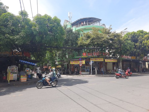 A sunny street corner lined with shops and trees, with motorbikes crossing the intersection