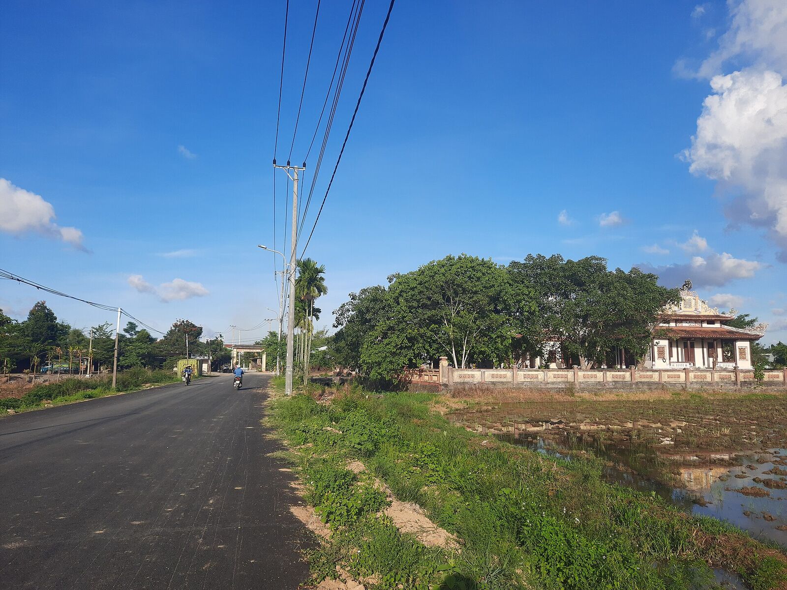 A newly asphalted village road beside a flooded paddy field and a traditional temple building in An Trạch, Hòa Tiến
