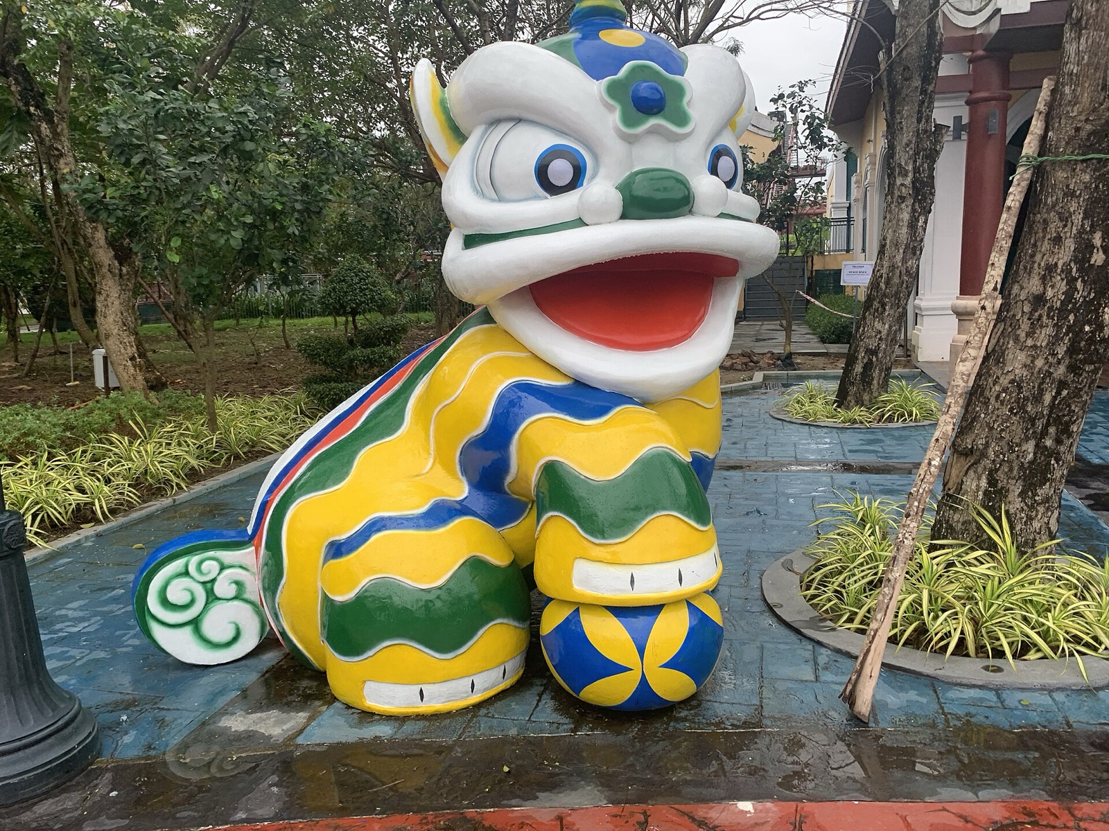A large, brightly painted lion statue in green, yellow and blue standing among plants and trees at an amusement park in Da Nang