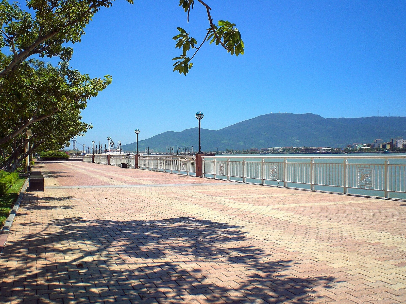 A paved riverside promenade with a white railing and lamp posts beside the Han River, with a green mountain across the water