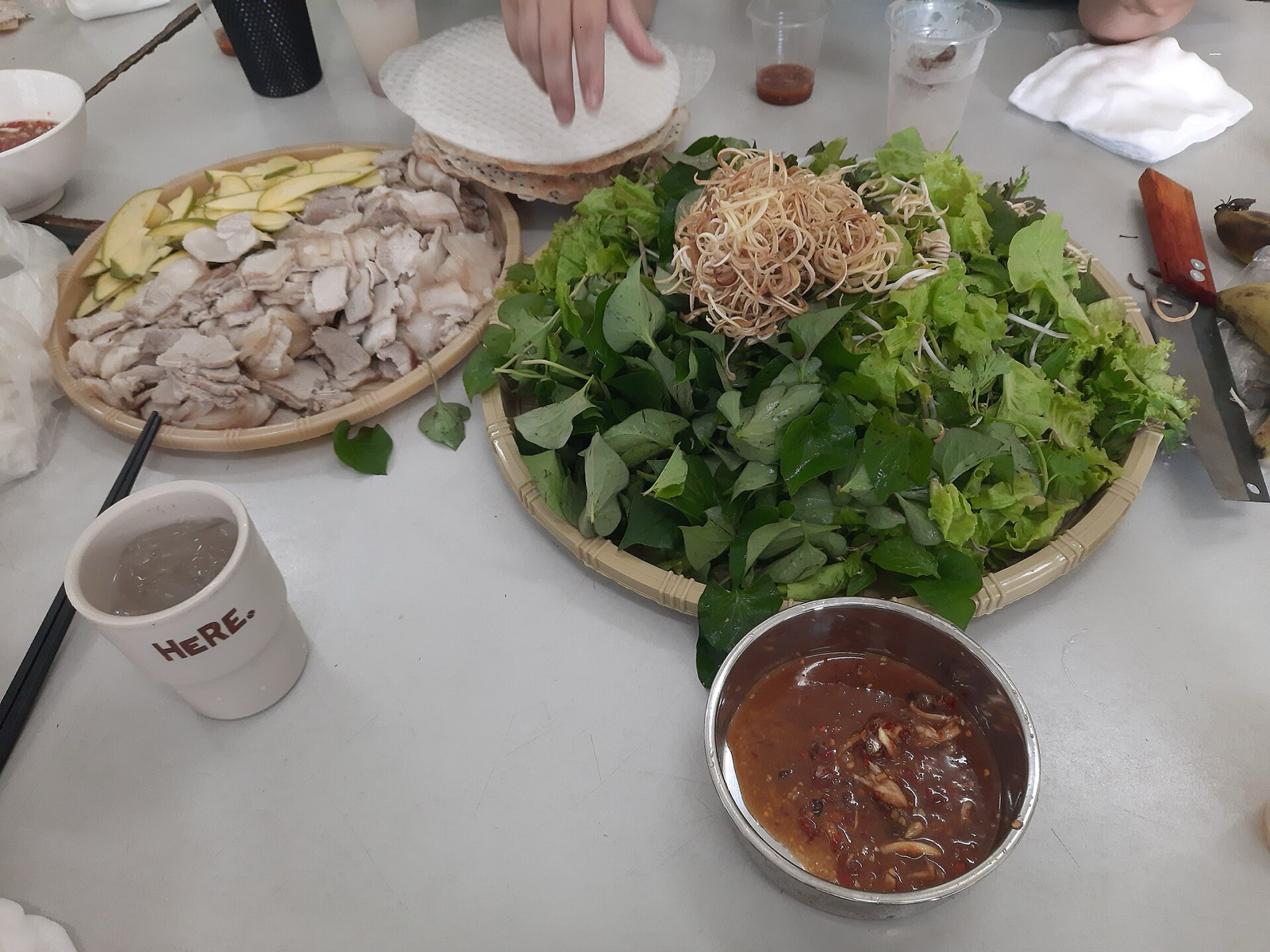 A table spread of thinly sliced boiled pork, a large plate of fresh herbs and greens, rice paper and a bowl of dark mam nem dipping sauce