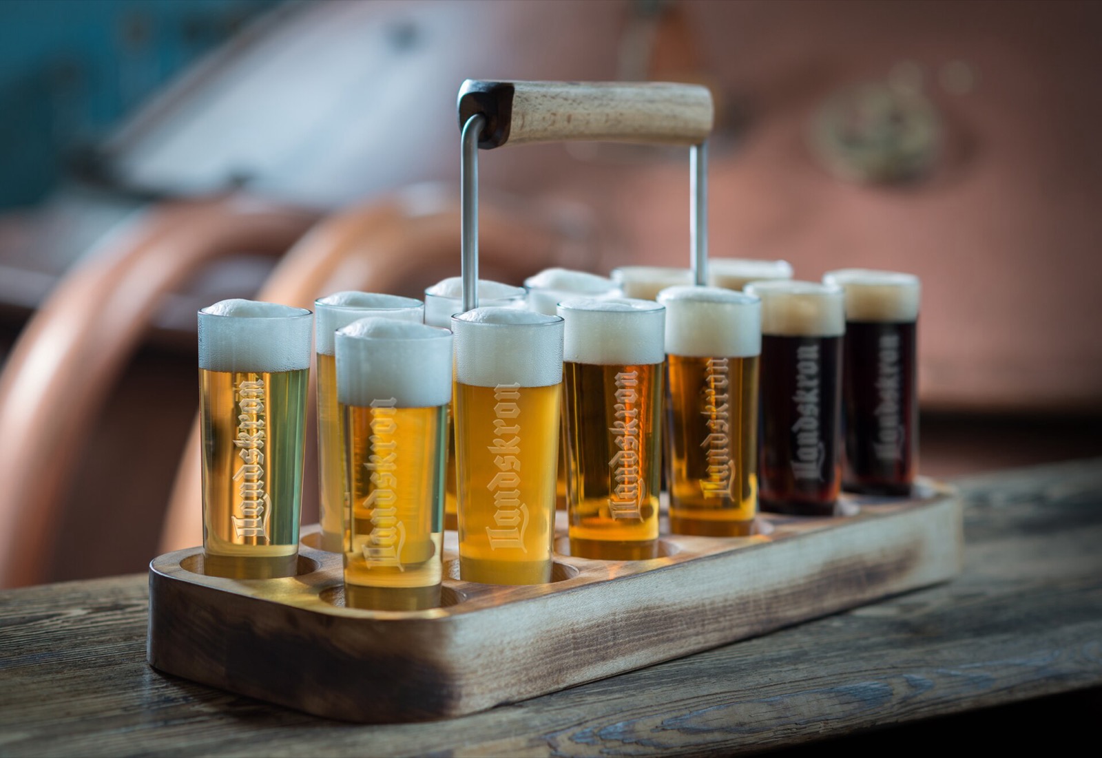 A wooden tasting tray holding a row of small glasses of beer in shades from pale gold to amber