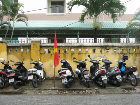 A row of parked motorbikes and scooters against a yellow wall on a Da Nang street, with a red flag above them