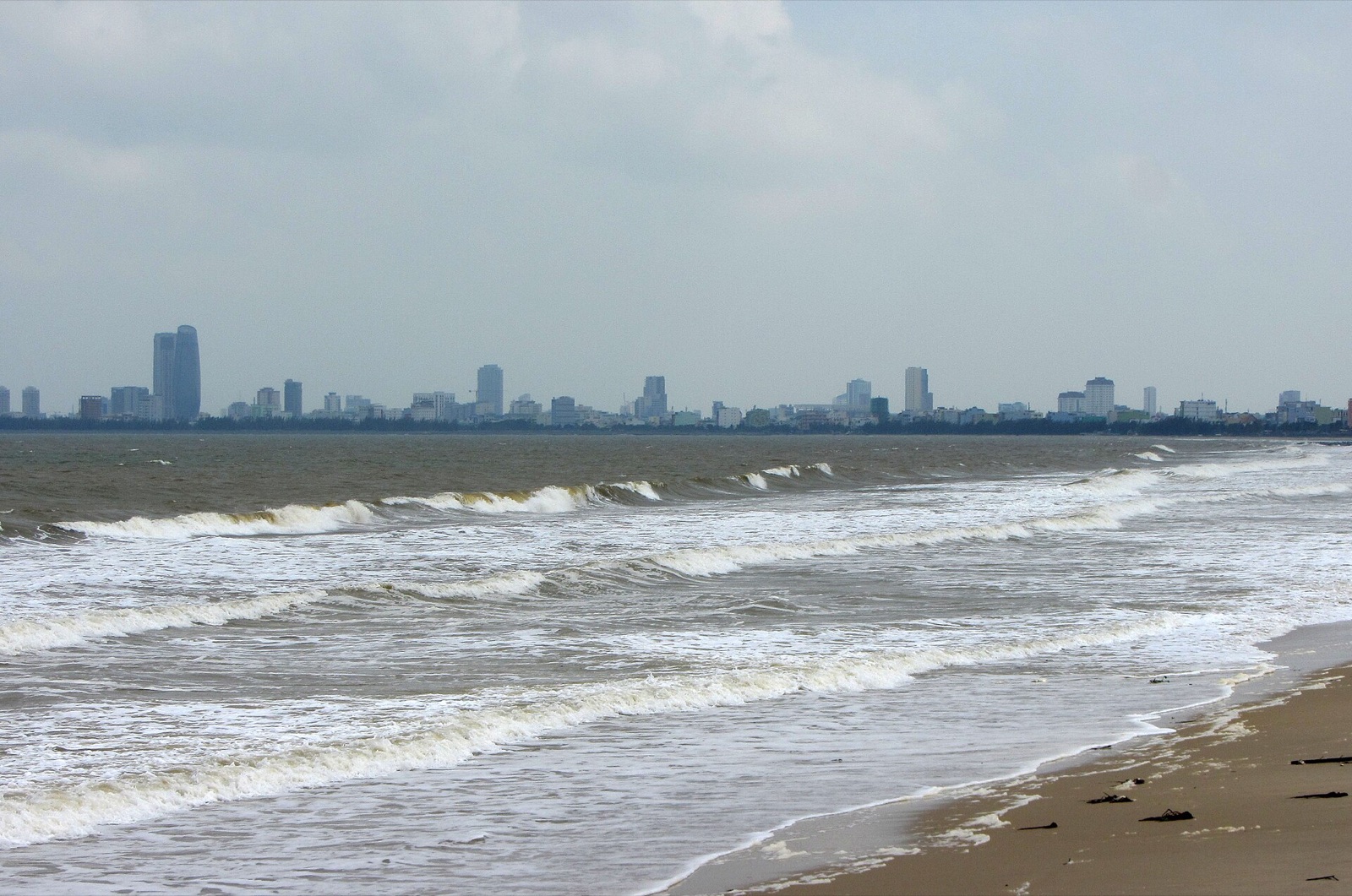 Grey waves breaking on a Da Nang beach under an overcast sky, with the city's high-rises hazy in the distance