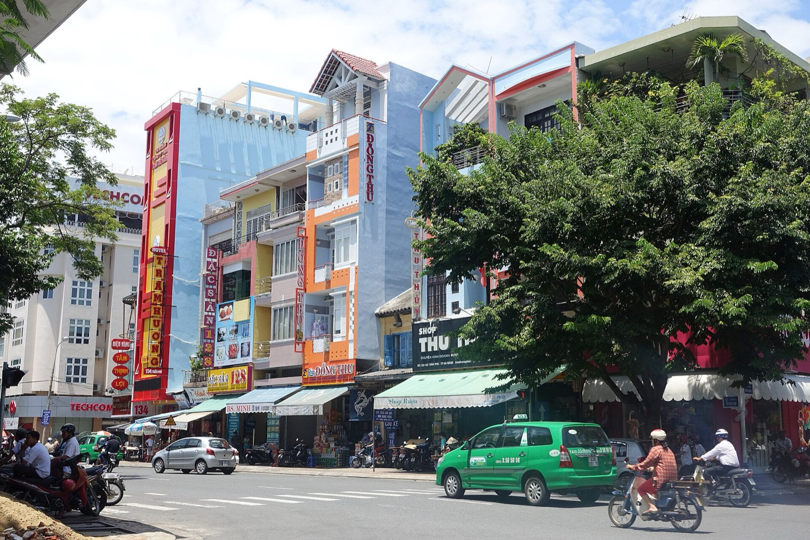 A busy Da Nang street with shophouses, signboards, trees, motorbikes and a green taxi