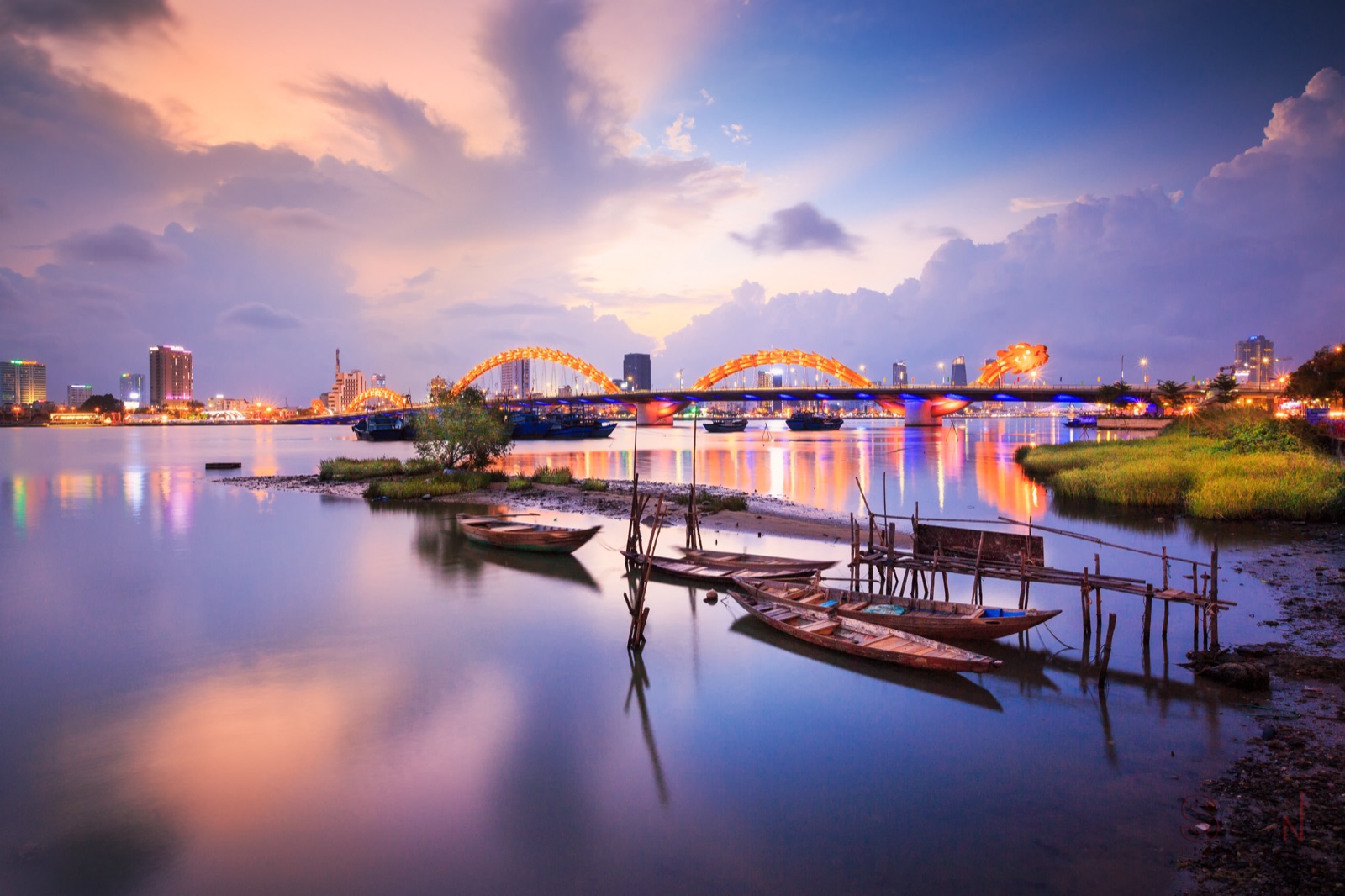 The Dragon Bridge and Da Nang skyline at dusk, reflected in calm water with small boats in the foreground