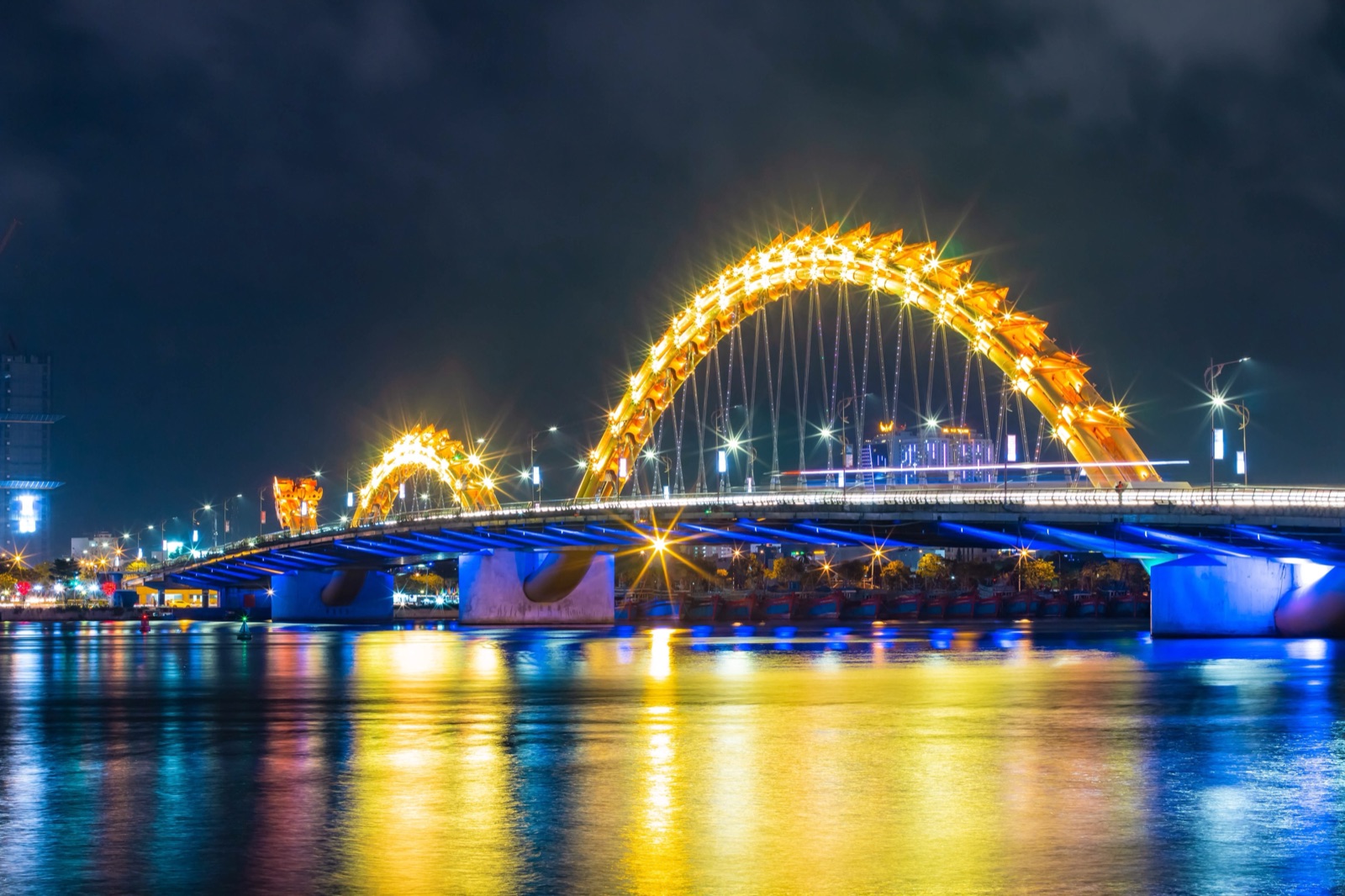 The Dragon Bridge lit up in gold and blue over the Han River at night, its arched dragon body reflected in the water