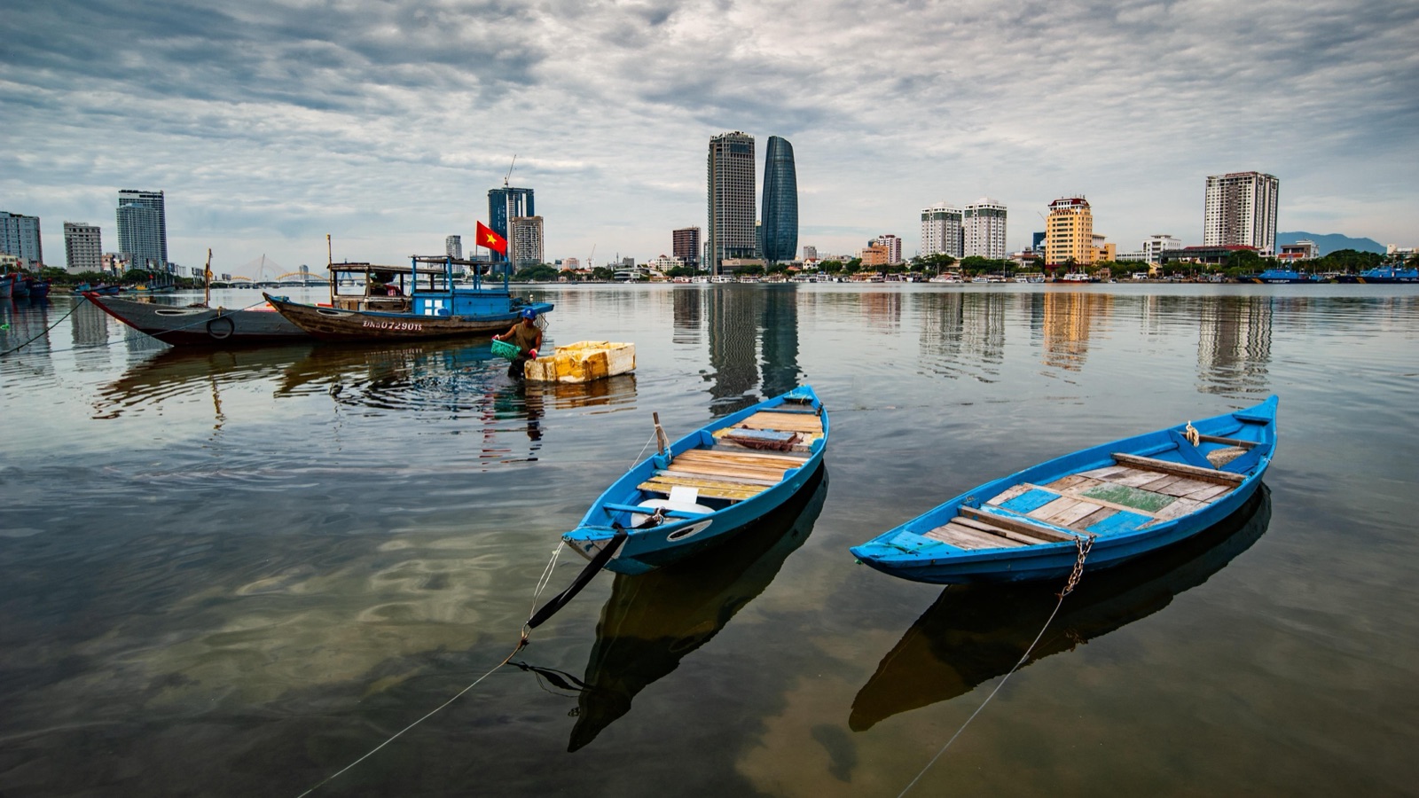 Two blue wooden boats on calm water in front of Da Nang's riverside high-rises