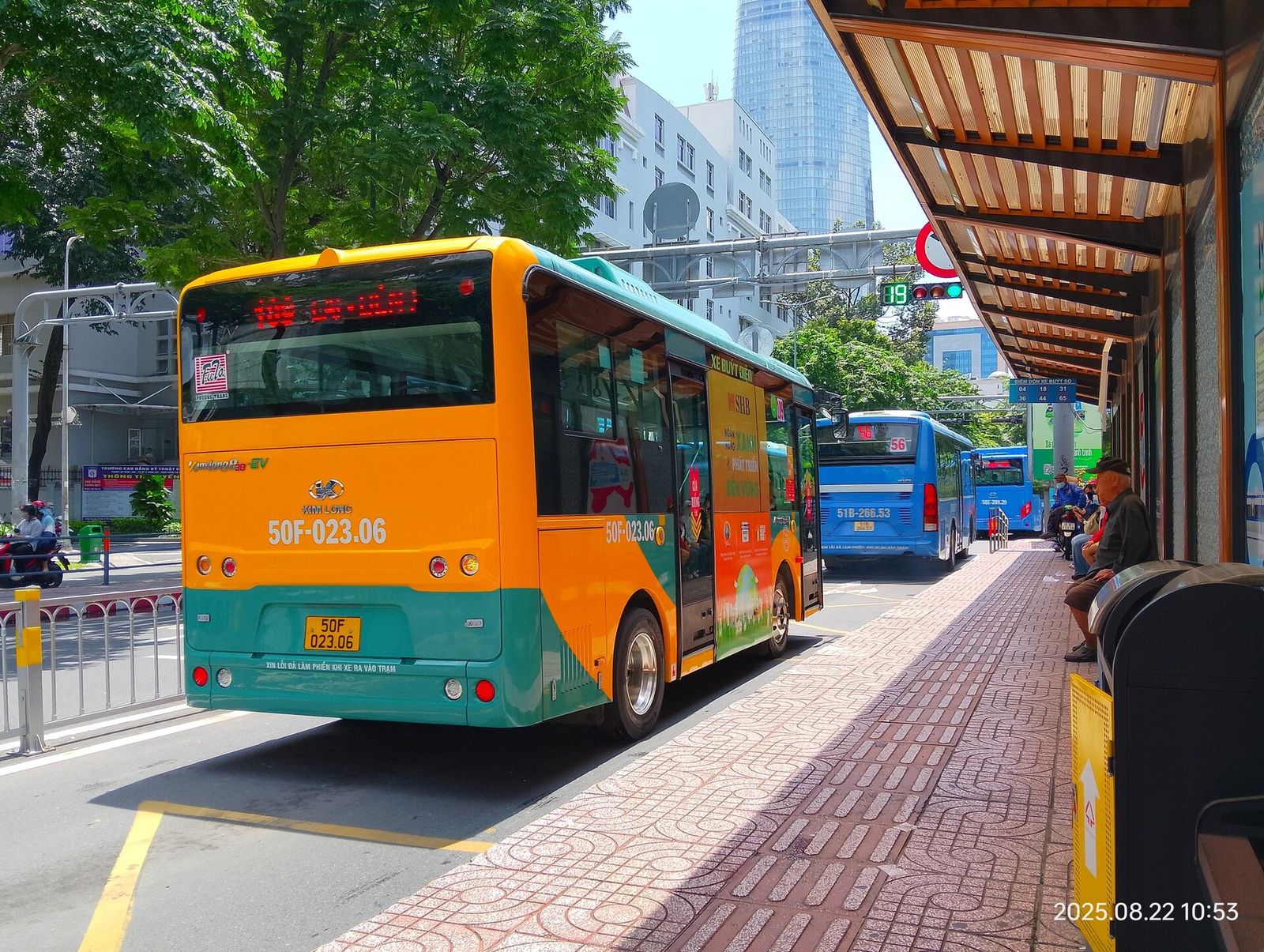 An orange and green city bus at a covered bus stop on a tree-lined street