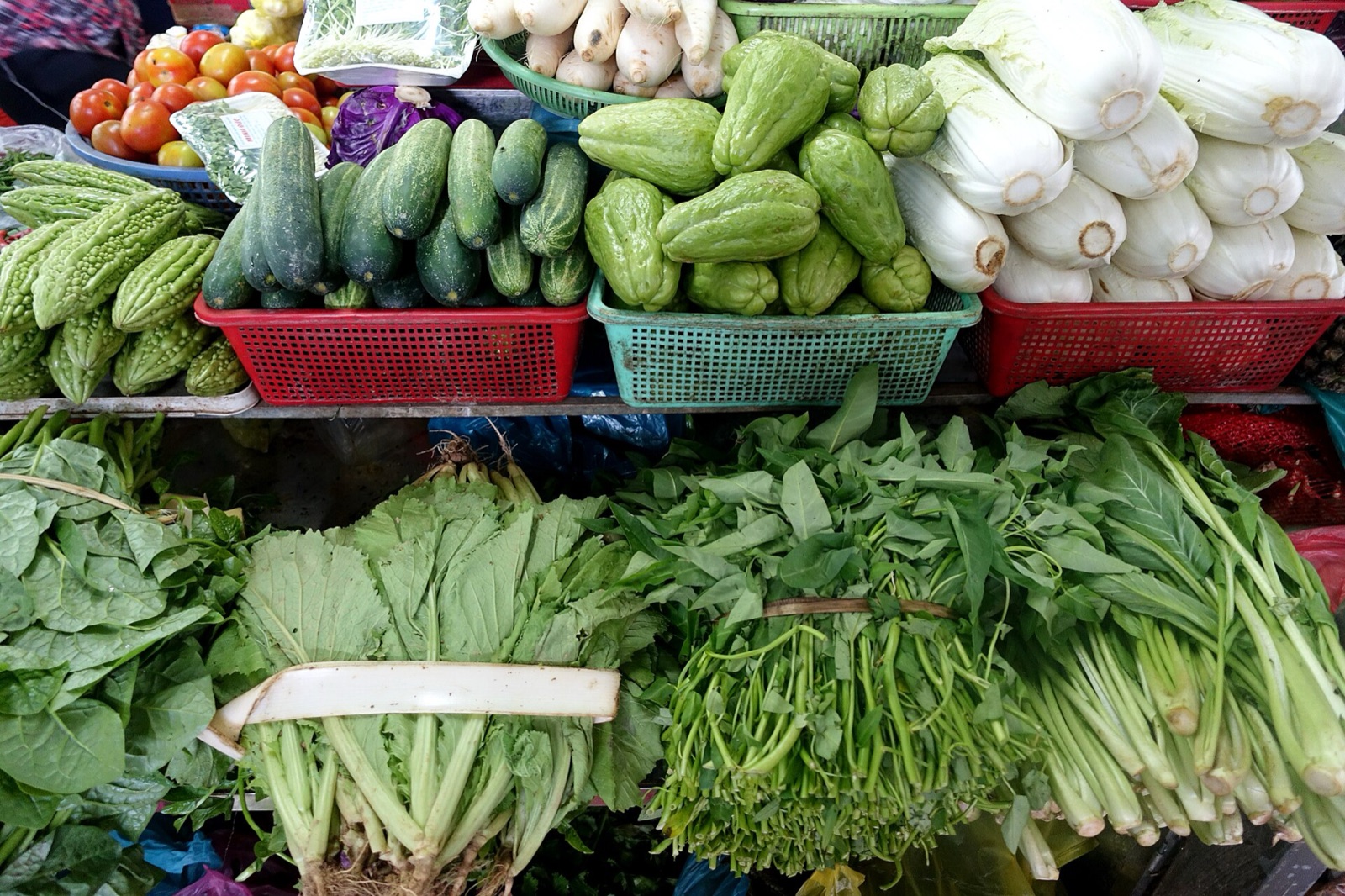 Baskets of cucumbers, bitter melon, cabbages and bundles of leafy greens stacked on a vegetable stall