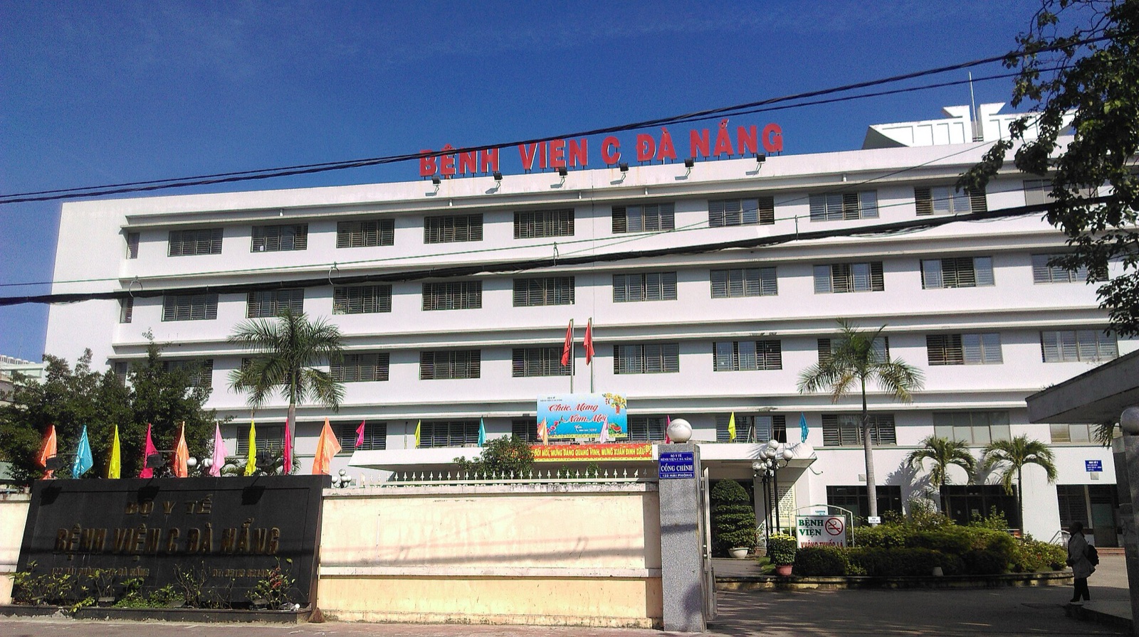 The white multi-story front building of Hospital C in Da Nang with its red name sign, palm trees and flags at the gate