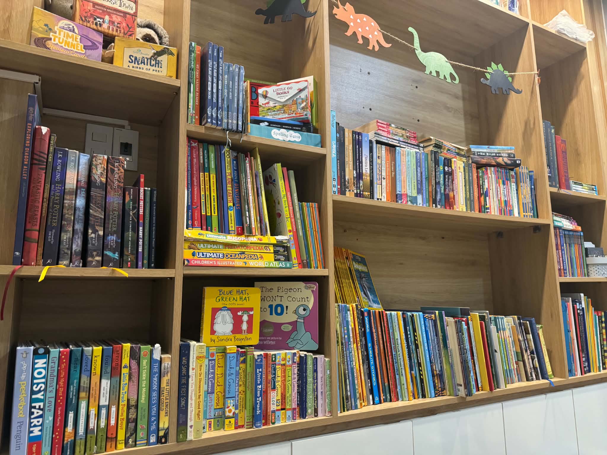 Wooden shelves of children’s books, games and puzzles with a paper dinosaur garland above