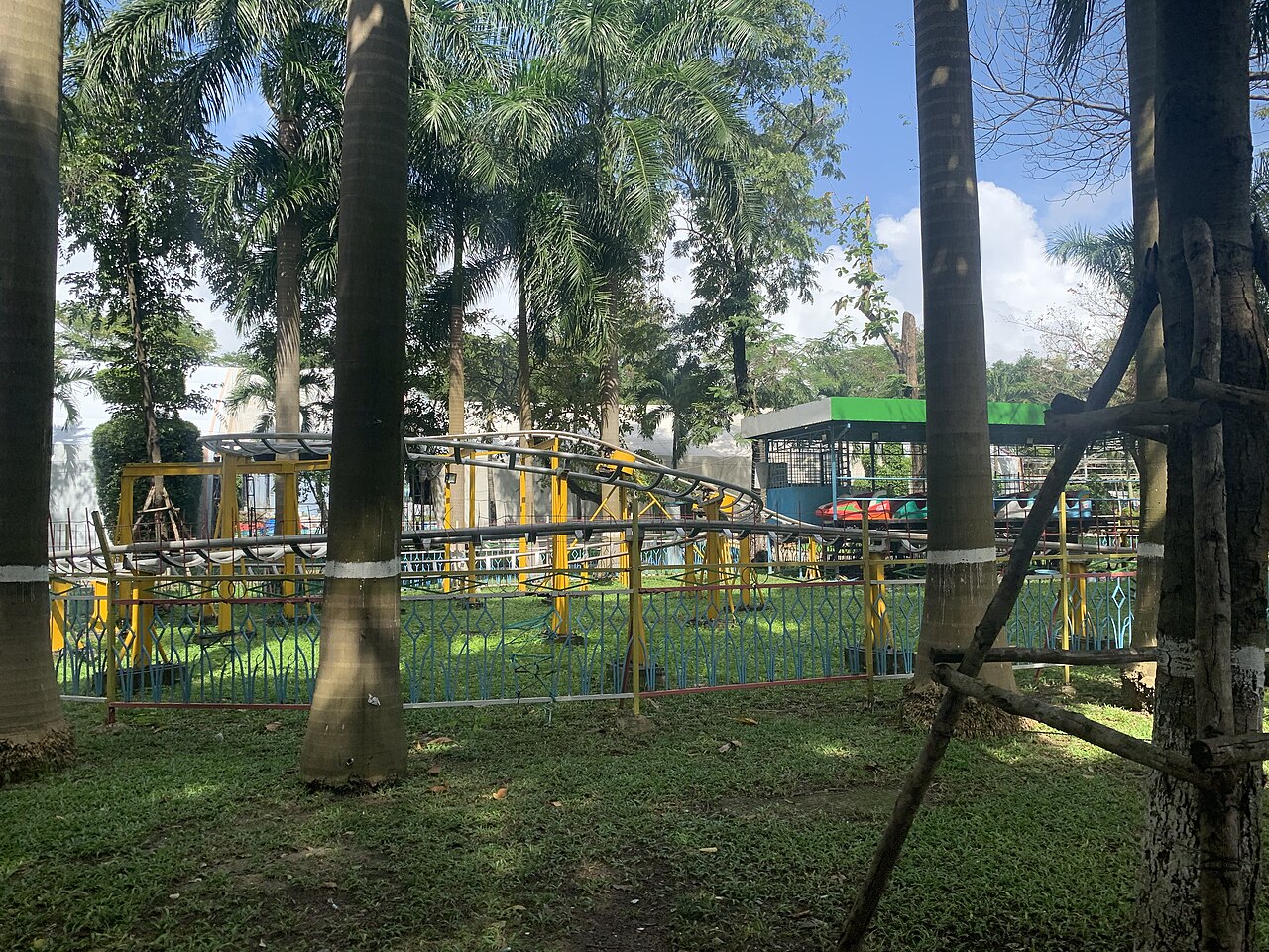 Palm trees and a small roller-coaster track behind a low fence on a grassy lawn in March 29 Park