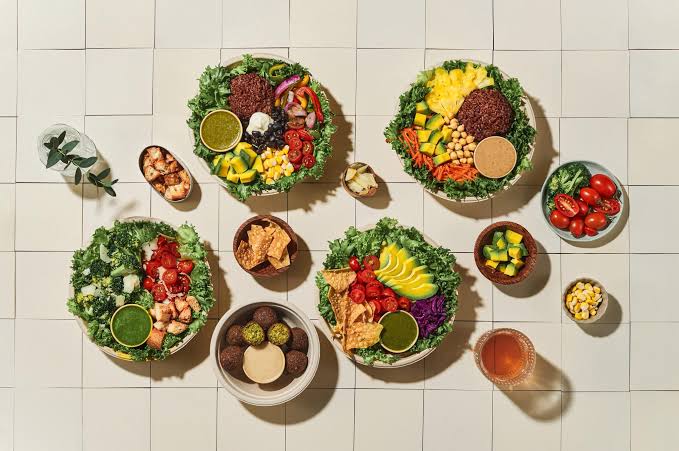 A top-down view of five salad and grain bowls with greens, tomatoes, avocado, corn and falafel, surrounded by small dishes of sauce, chips and a drink on a tiled surface