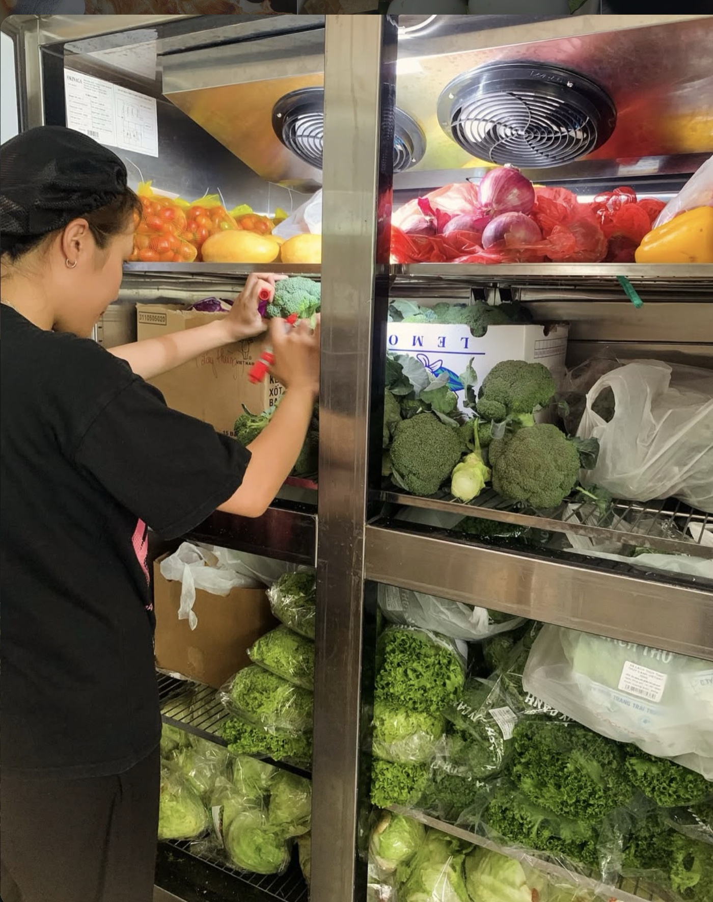 A kitchen worker in a black cap and shirt reaches into a steel refrigerator stocked with broccoli, lettuce, tomatoes, onions and peppers