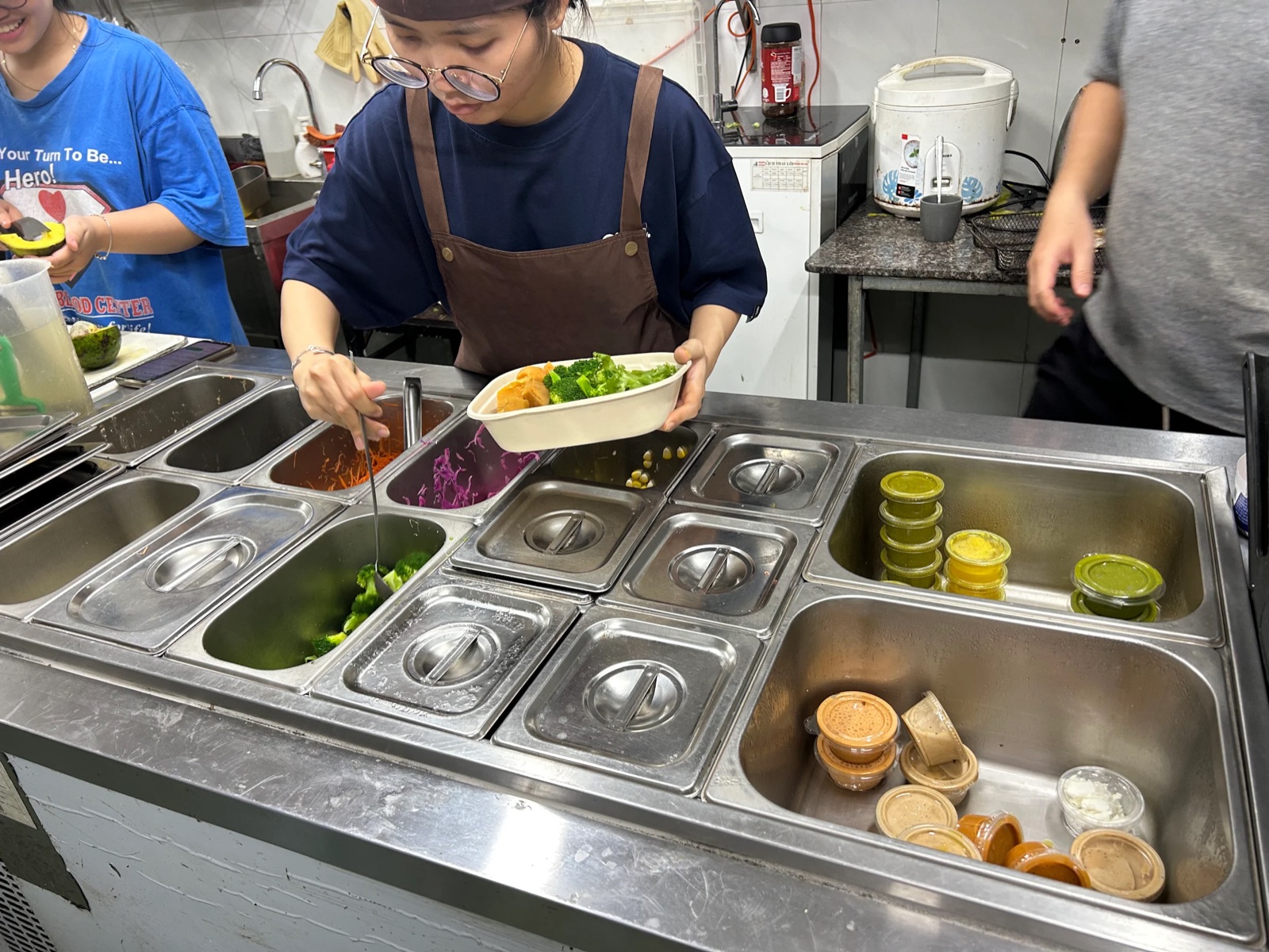 A kitchen worker in glasses and a brown apron holds a bowl of broccoli and pumpkin over a counter of steel containers holding salad ingredients and sauce cups