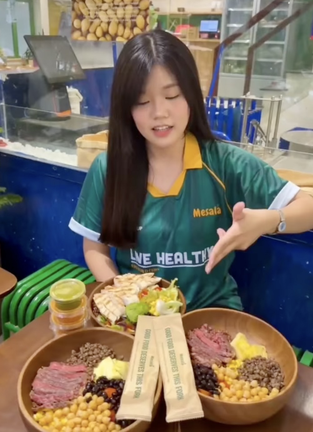 A woman in a green Mesala jersey sits behind two wooden bowls of beef, chicken, lentils, chickpeas and black beans, with small cups of sauce beside them