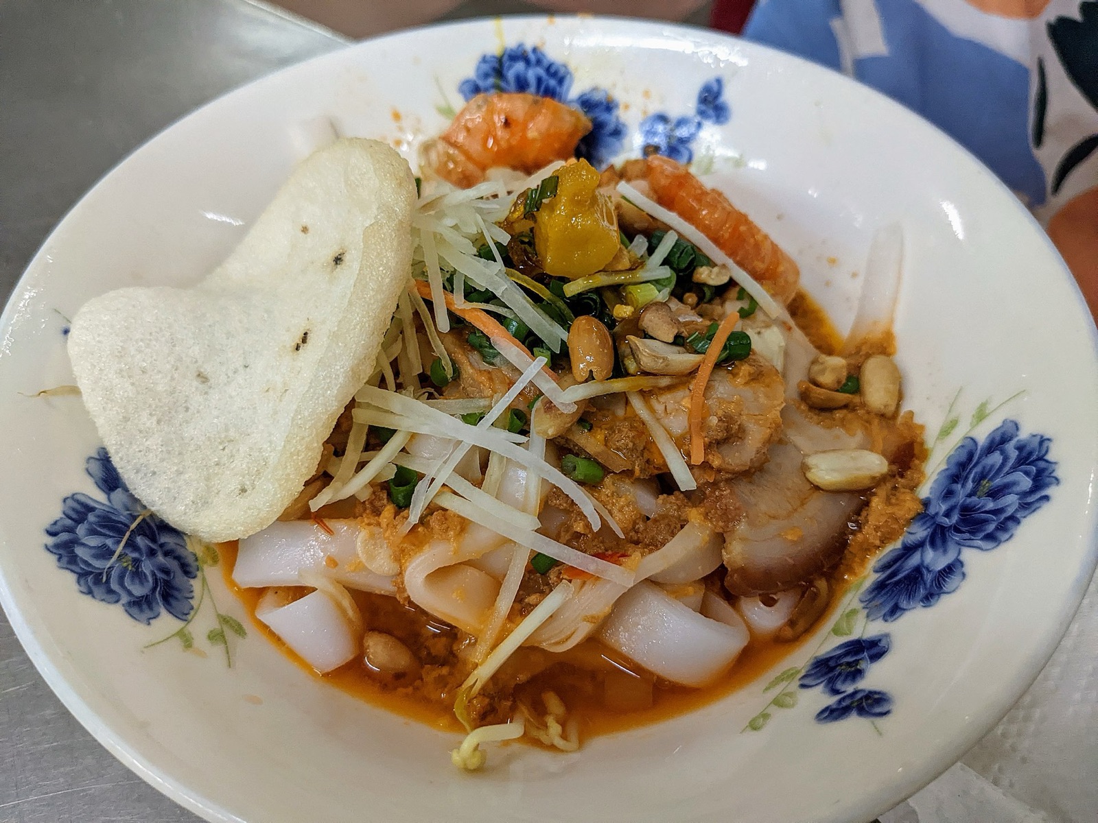 A bowl of mi Quang with wide yellow noodles, shrimp, pork, quail egg and herbs, a rice cracker on the side, in a blue-and-white bowl