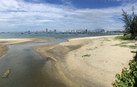 A wide sandy beach and a tidal stream with fishing boats in the bay and a high-rise skyline across the water
