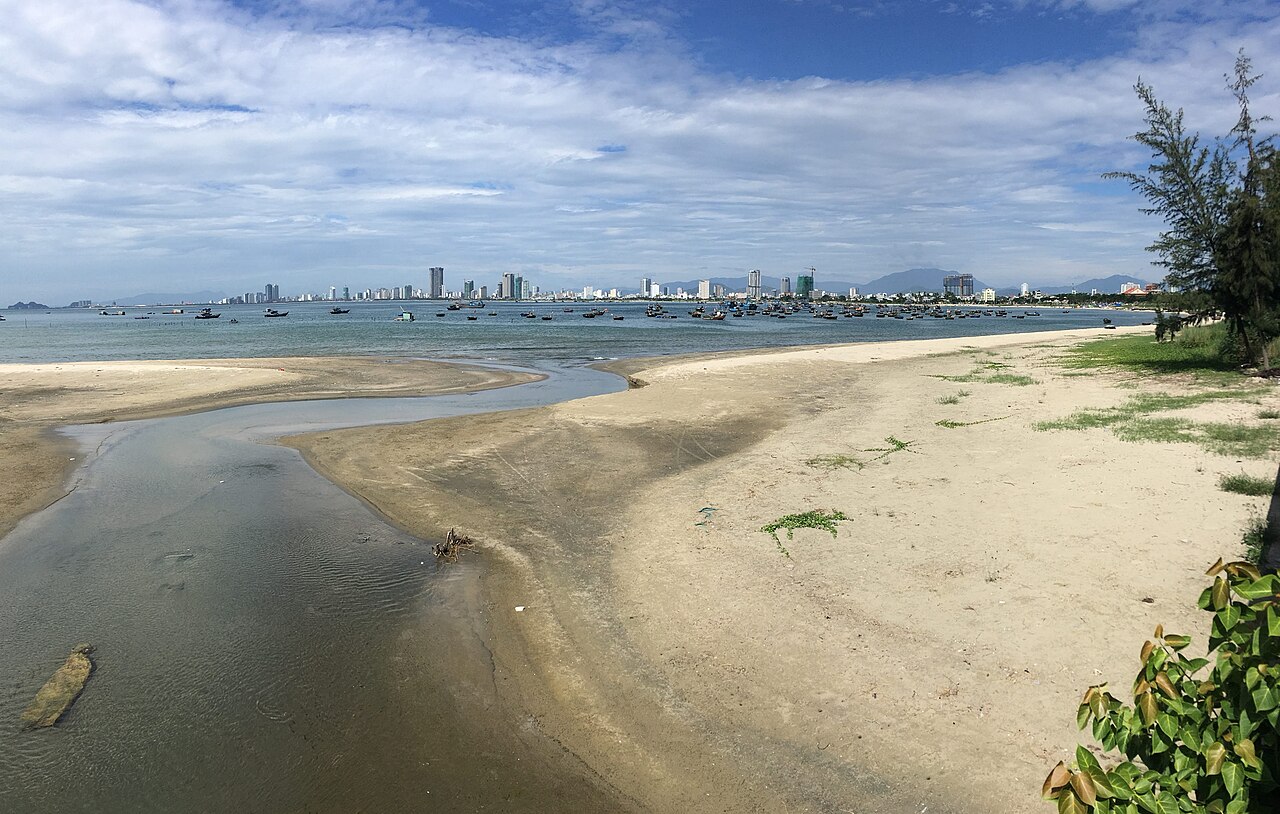 A wide sandy beach and a tidal stream with fishing boats in the bay and a high-rise skyline across the water