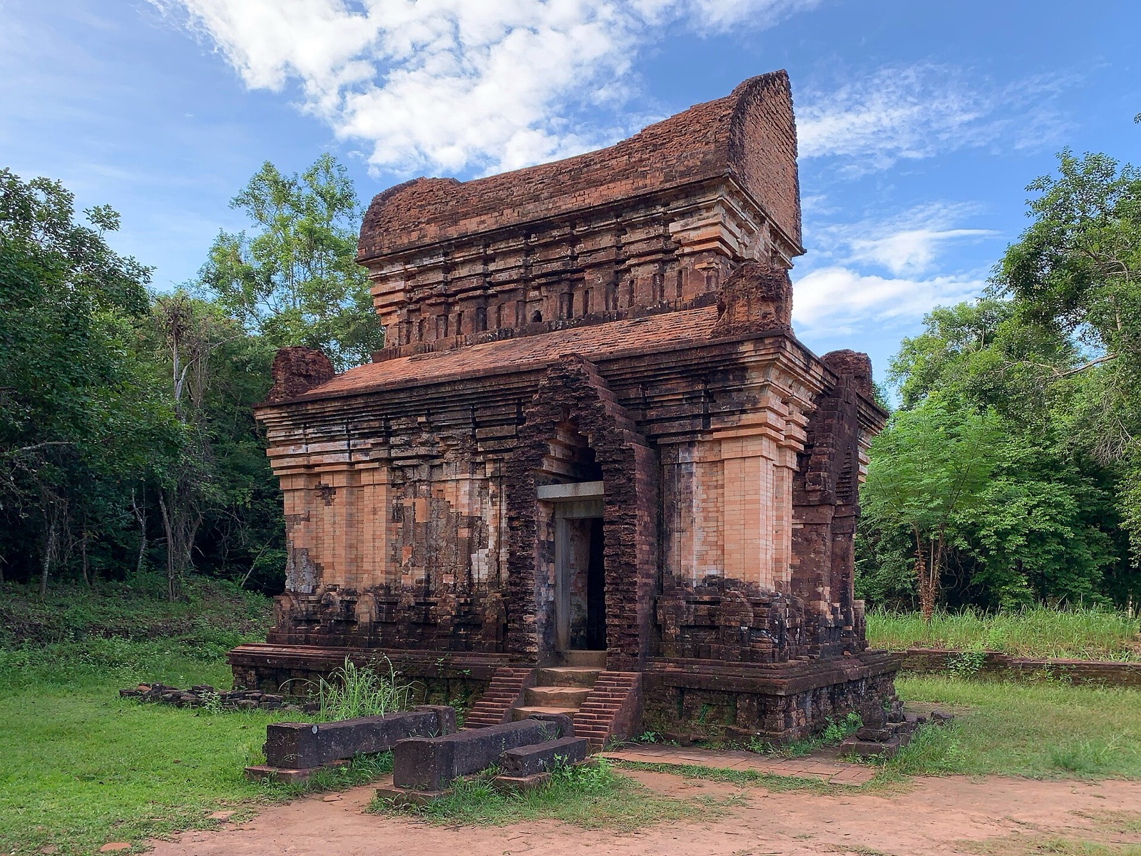 A small red-brick Cham temple with carved walls and a curved roof standing among trees and grass at My Son Sanctuary