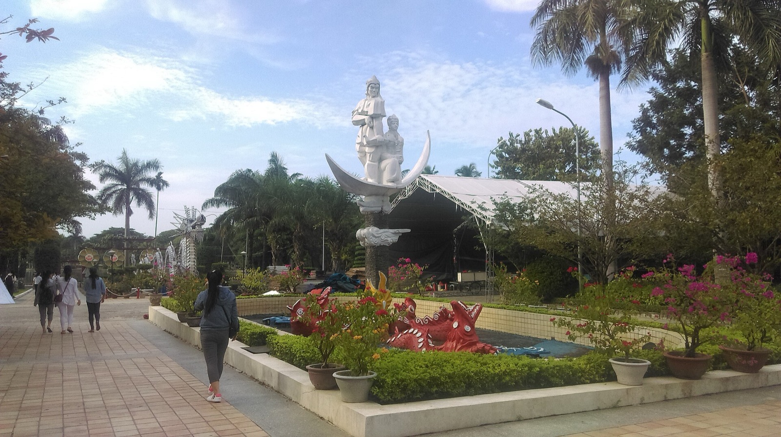 A paved path through flower beds and palm trees at 29/3 Park in Da Nang, with a white monument in the background and a few people walking